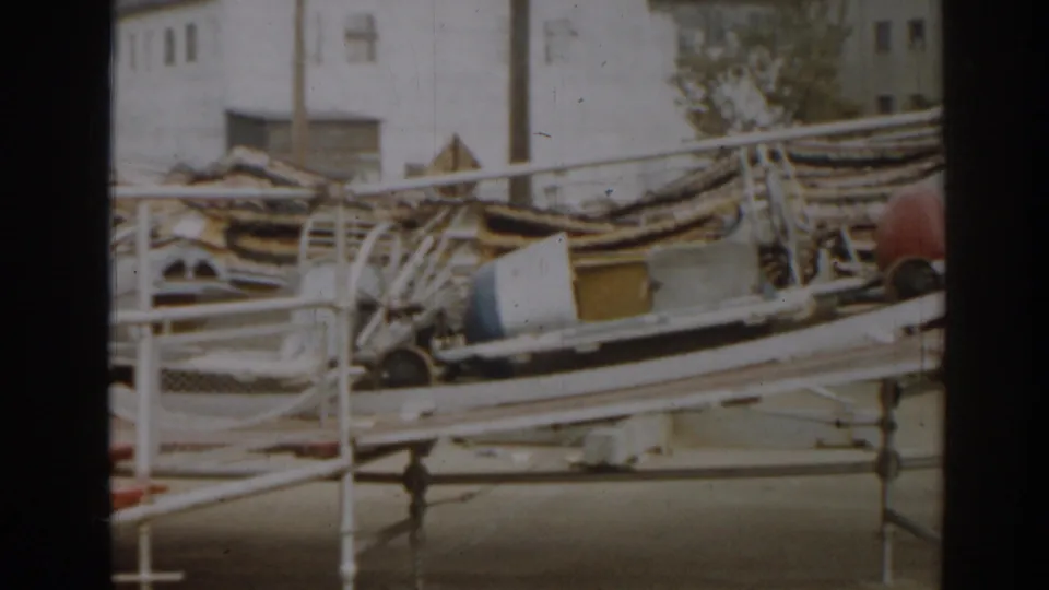1963 New Jersey 8mm frame with wrecked boats, twisted railings, scattered debris, and a waterfront building behind them.