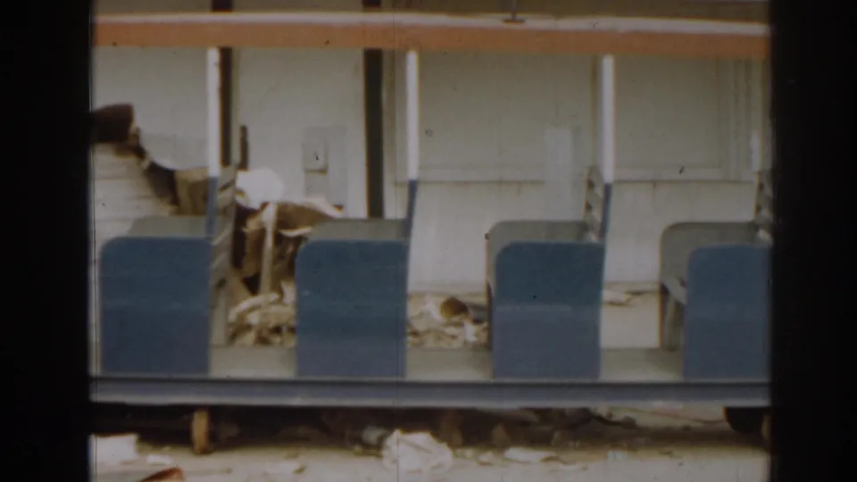1963 home-movie frame of a damaged miniature train car, emphasizing blue seats, pale supports, and the open interior area.