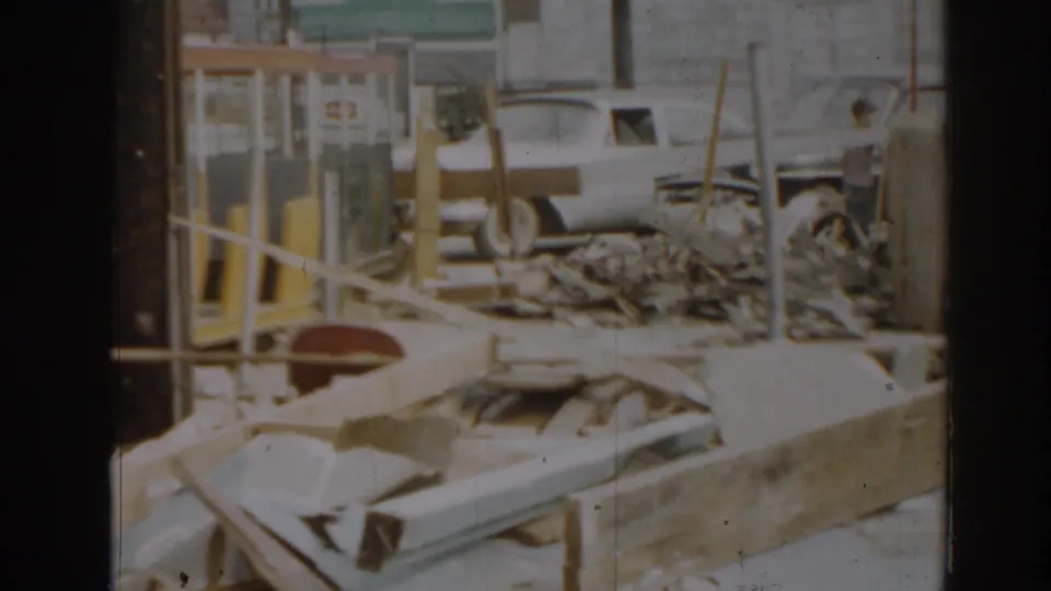 1963 New Jersey 8mm frame showing debris piles, railings, yellow posts, and a light-colored vehicle near damaged structures.