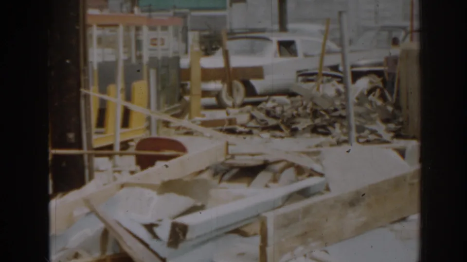 1963 New Jersey home-movie frame showing scattered wreckage, yellow posts, railings, and a light-colored vehicle outside damaged buildings.