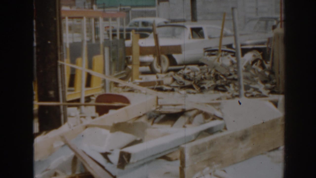 1963 New Jersey home-movie frame showing scattered construction debris, wooden rails, and a white period car beyond the site.