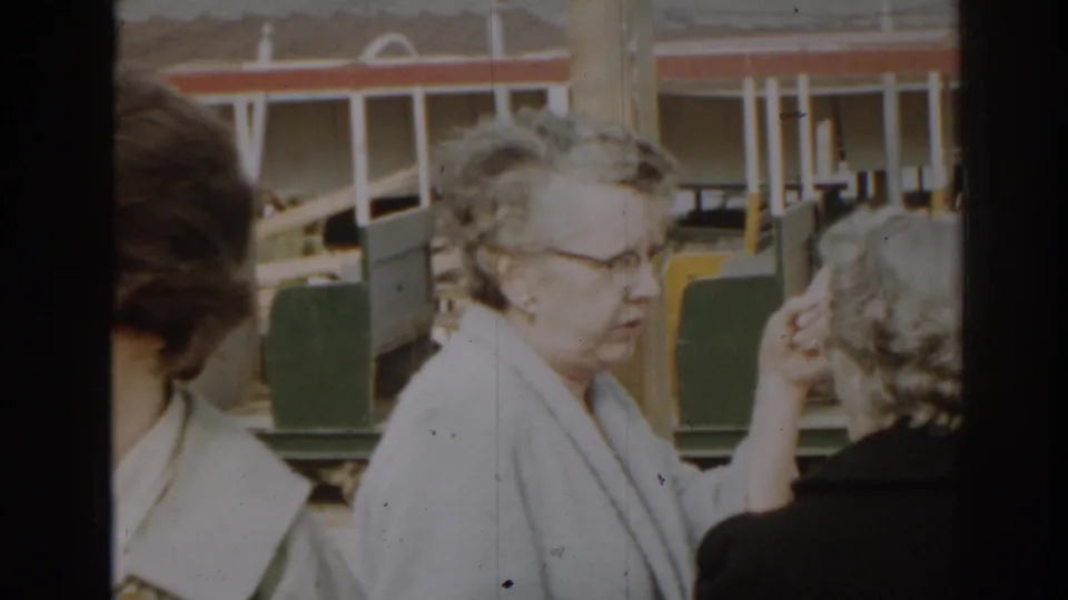 1963 New Jersey home-movie frame with an older woman in glasses and light coat talking near carts and a building-site backdrop.