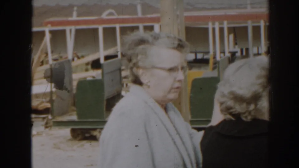 1963 New Jersey frame showing women in conversation near green carts, posts, and a partially visible building-site structure.
