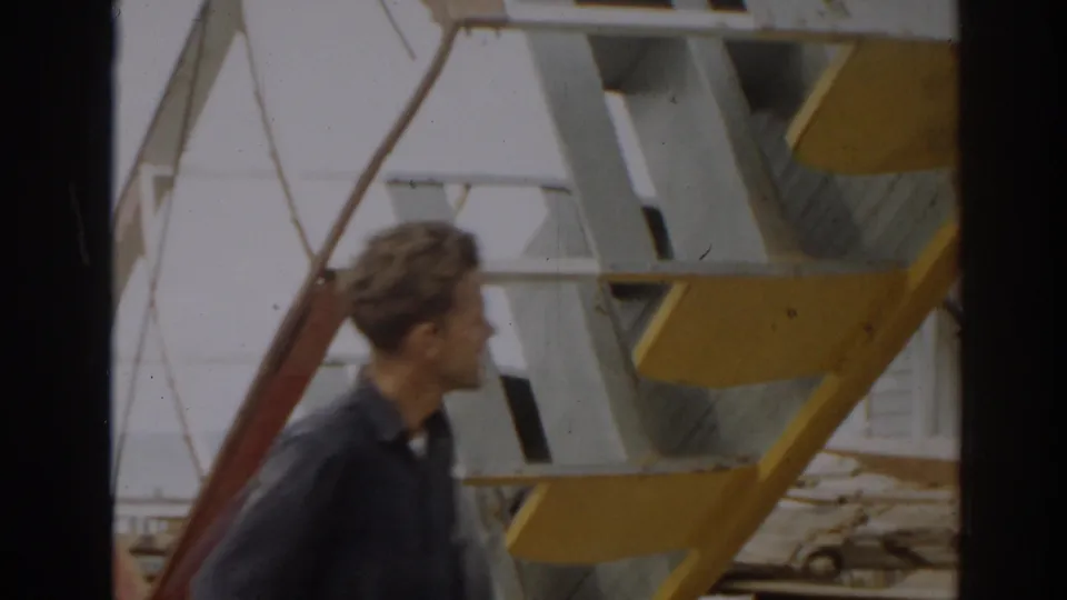 A man passes through a debris-strewn 1963 New Jersey scene beside yellow and white metal framework and scattered boards.