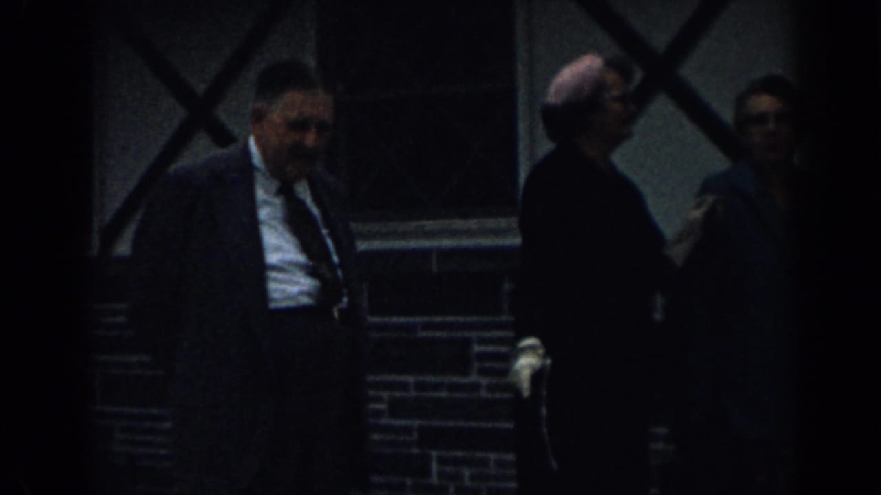 Man in a suit and other adults walk beside a brick wall and window.
