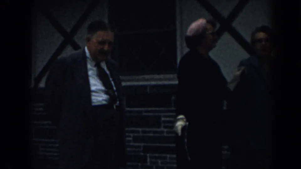 A man in a dark suit and tie walks near other adults beside a brick wall and cross-braced window in 1963 New Jersey footage.