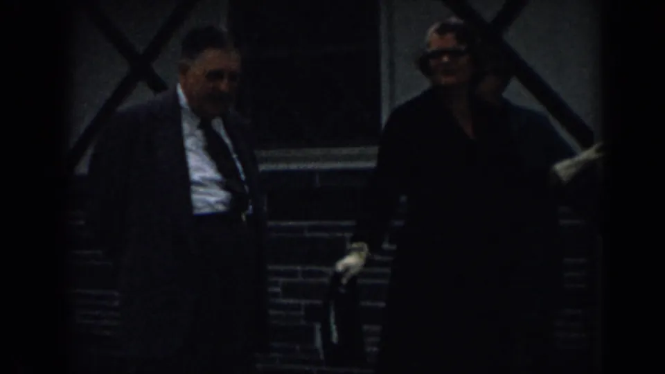 A suited man walks beside a woman wearing glasses and a dark coat near a brick building in the 1963 New Jersey home-movie clip.