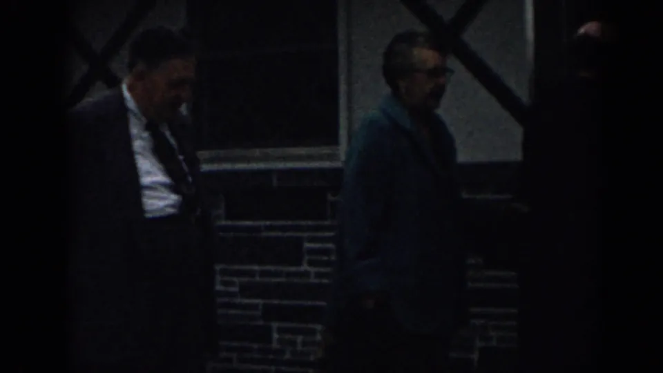 The man in a suit and the woman in a dark coat move past the brick wall and cross-braced window, partly covered by low light.