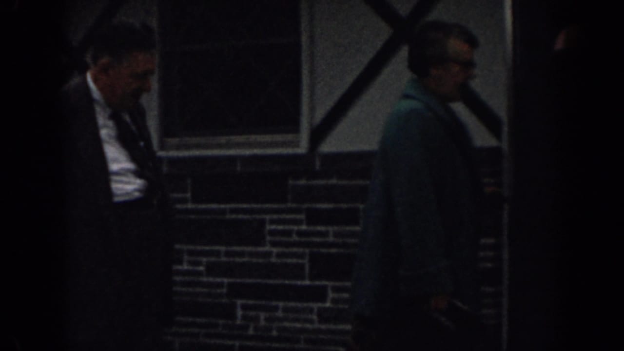 Two adults walk past a dark doorway and cross-braced window on a masonry wall.