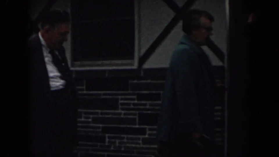 In 1963 New Jersey 8mm footage, two adults pass a dark doorway beside a masonry wall and window with black X-bracing.