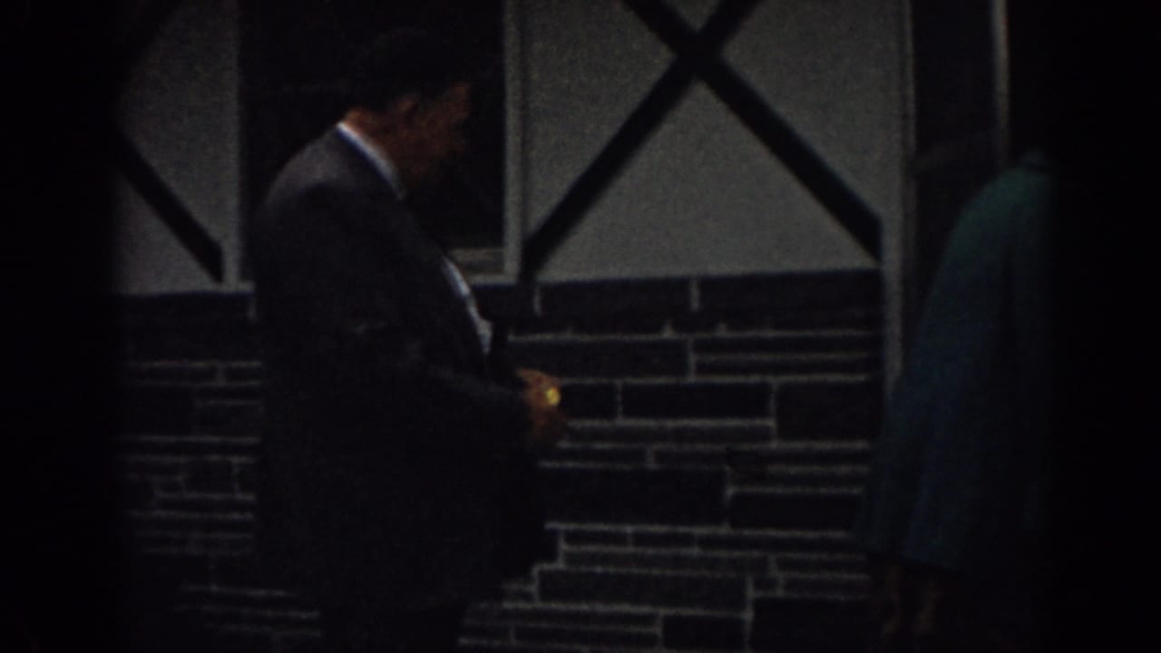A suited man stands near a dark doorway while another adult in teal is partly visible.