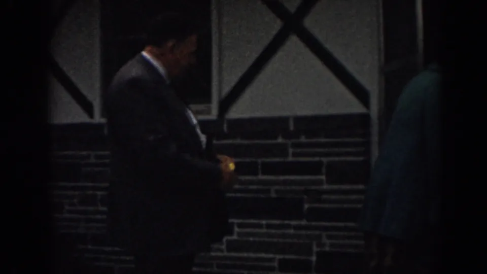 A suited man appears beside a shadowed entrance, with a teal-jacketed adult partly visible against the cross-braced wall.