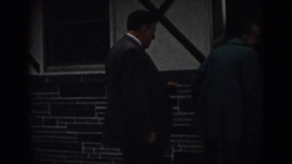 The adults continue along the building front in 1963 New Jersey footage, partly obscured by shadow and soft focus.