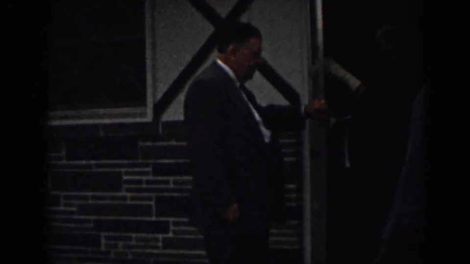 A man in a dark suit nears a doorway in the dim 8mm frame, with another adult in teal partly visible at right.
