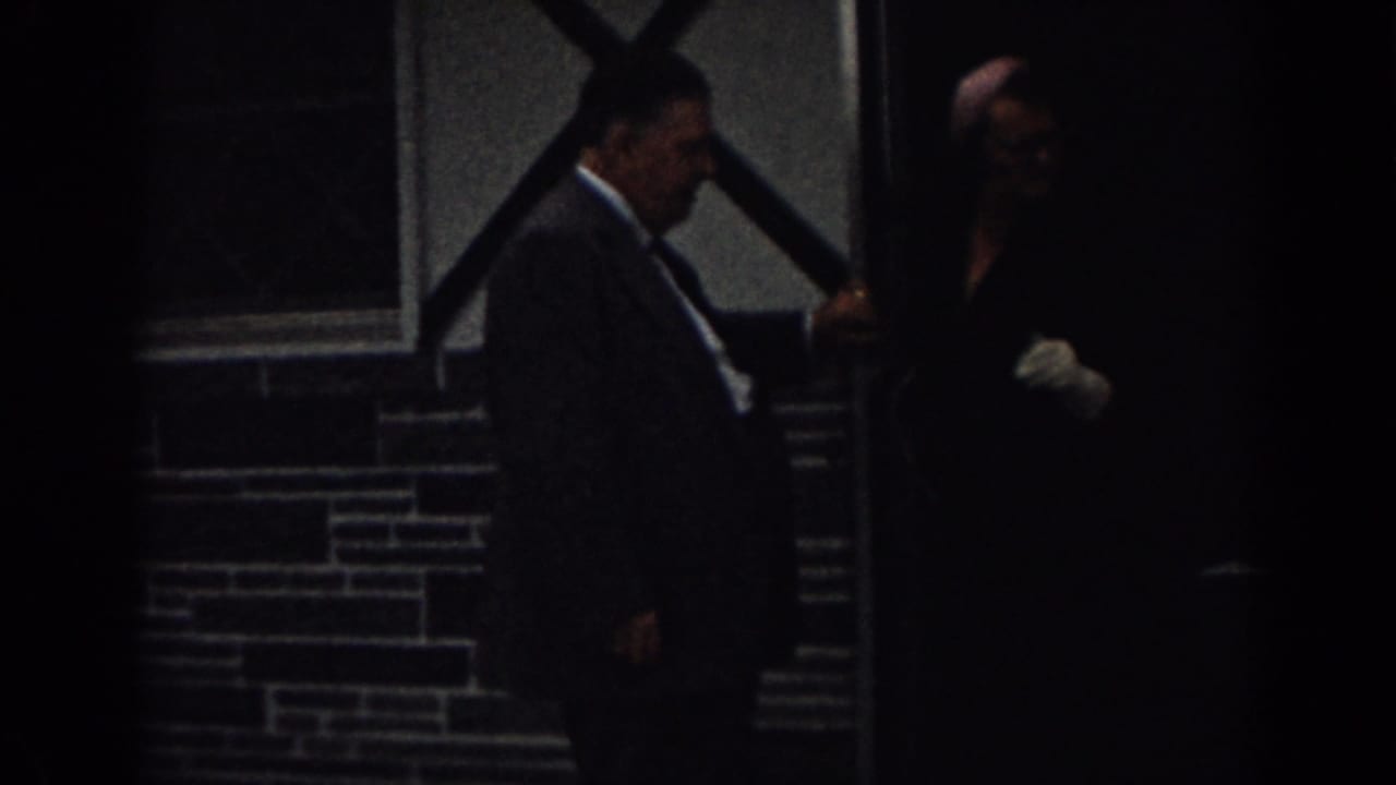 1963 New Jersey 8mm frame of a suited man at a dark doorway with a woman in dark clothing and white gloves nearby.