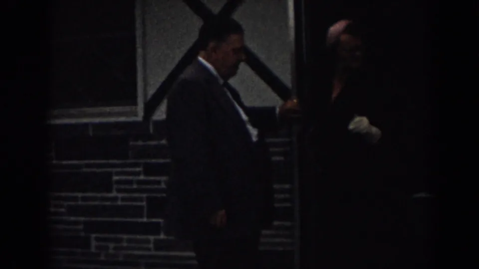 1963 New Jersey 8mm frame of a suited man at a dark doorway with a woman in dark clothing and white gloves nearby.