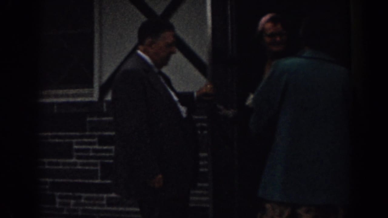A suited man holds the door as a person in a teal coat approaches or enters.