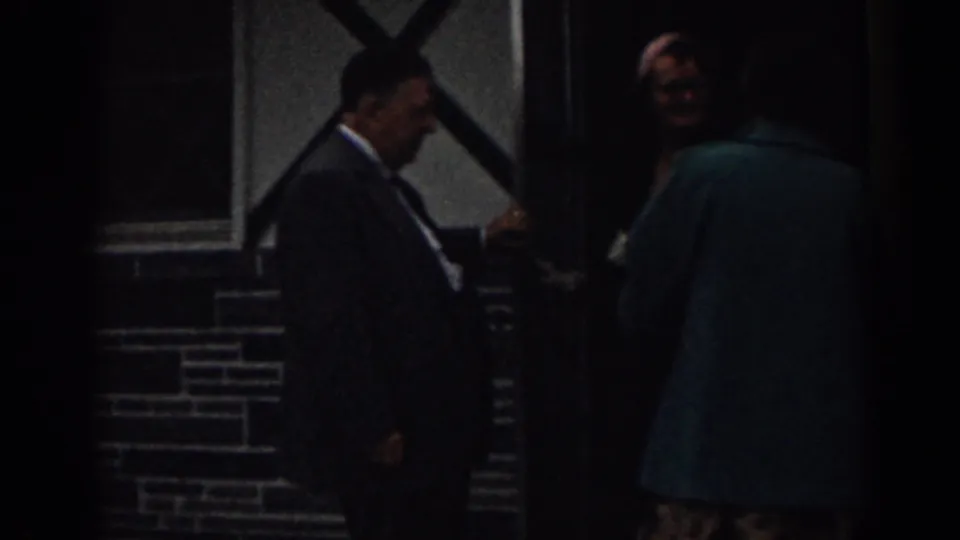 1963 New Jersey home-movie frame showing a man in a suit holding a dark entrance door for a person in a teal coat.