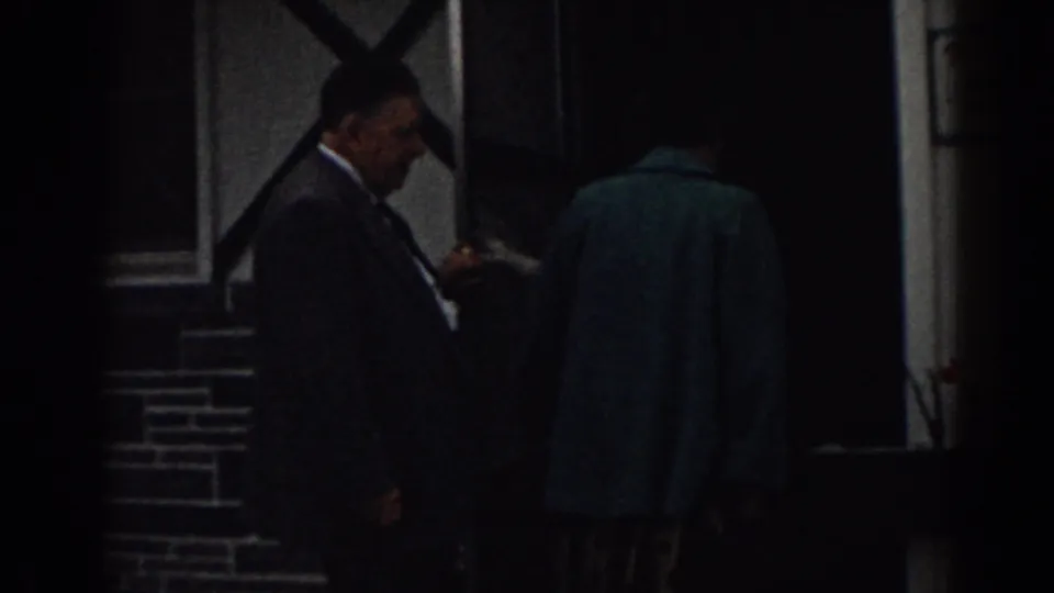 Low-light 1963 New Jersey 8mm frame with a suited man beside a brick entrance and an adult in a teal coat entering.