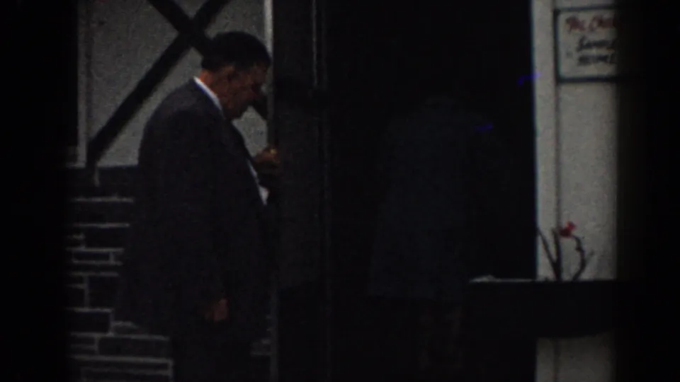 1963 New Jersey 8mm frame of a suited man near a shadowed building entrance, with brickwork and a small wall sign visible.