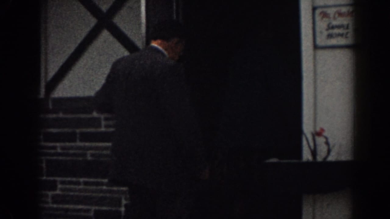 Man in a dark suit entering a doorway beside a stone-faced house.