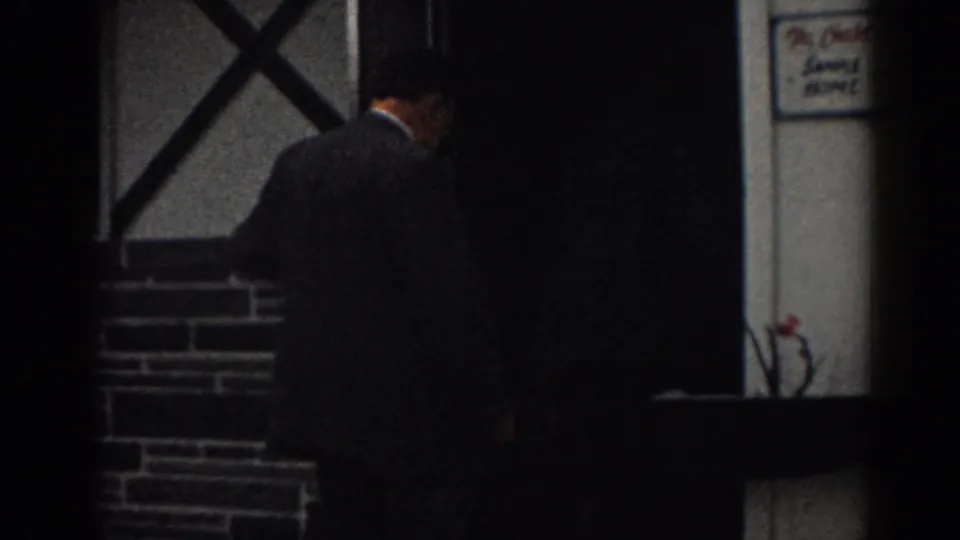 In 1963 New Jersey footage, a man in a dark suit appears near a doorway on a stone-faced residential exterior.