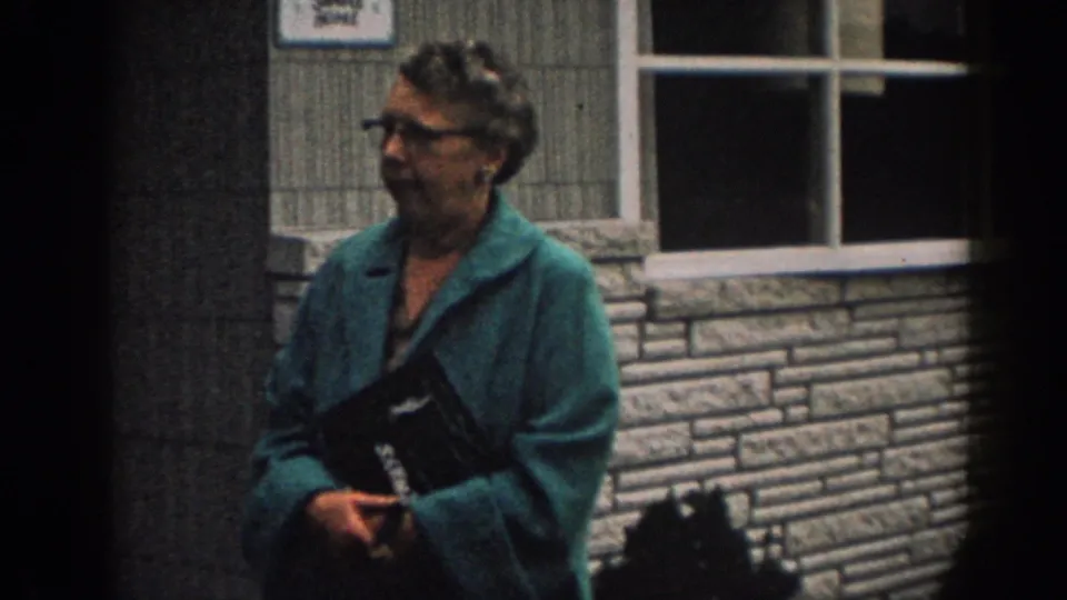 In 1963 New Jersey home-movie footage, a woman in a teal jacket and glasses stands near a window and stone-facing.