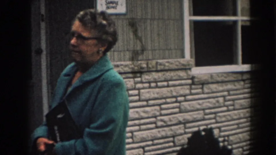 In 1963 New Jersey footage, the woman walks in profile past the stone-faced house while carrying a dark item.
