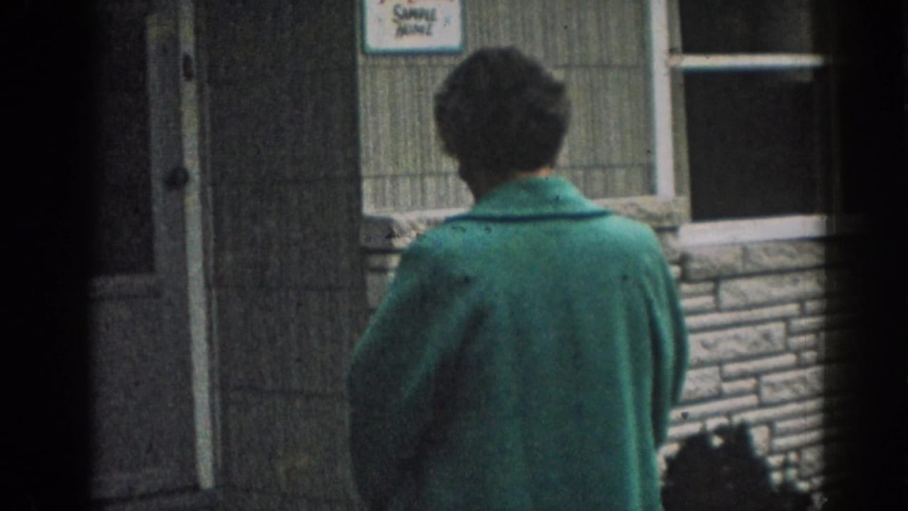 Woman in a green coat stands outside a building, seen from behind.