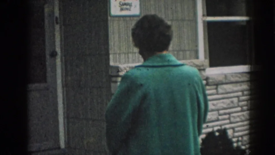 In 1963 New Jersey home-movie footage, a woman in a green coat stands with her back to the camera outside a small building.