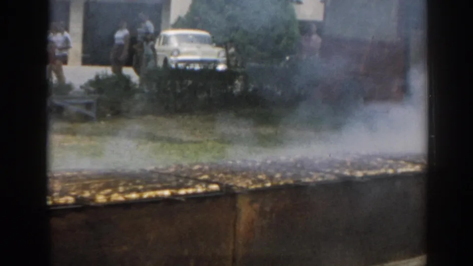 1963 New Jersey 8mm frame showing smoke rising from a large outdoor grill, with people and a white car beyond the yard.