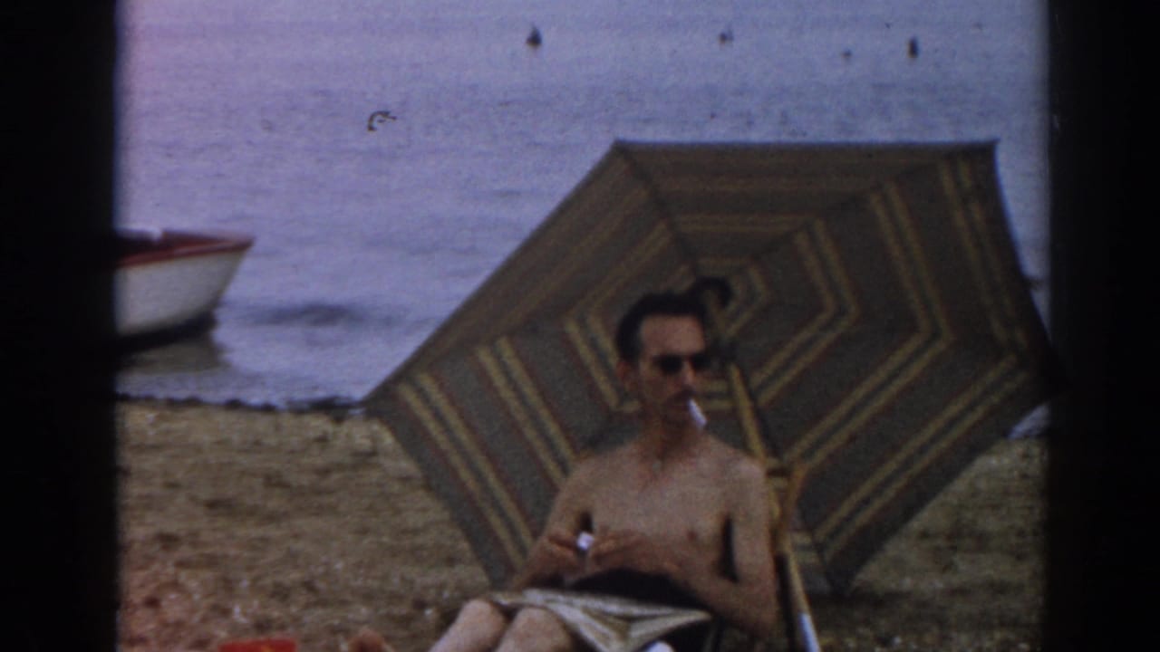 Shirtless man seated under a striped beach umbrella near the water.