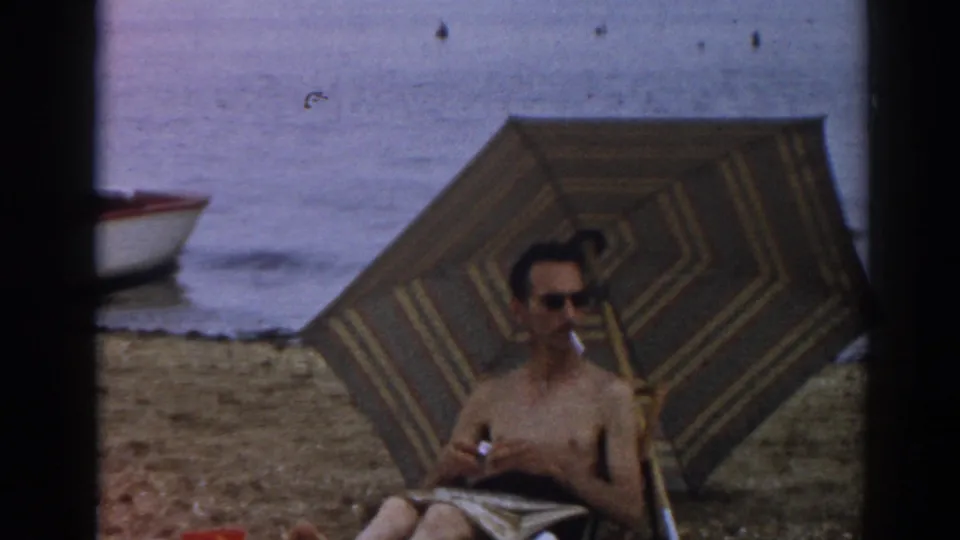 In 1963 New Jersey home-movie footage, a shirtless man sits under a striped beach umbrella with a cigarette near the shoreline.