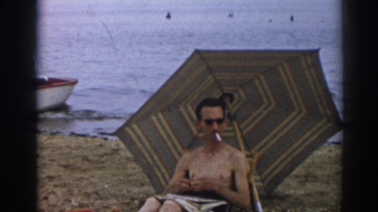 Man holding a cigarette while seated beneath a striped umbrella at the beach.