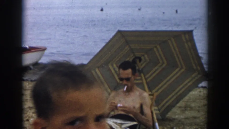 A blurred head crosses the foreground while the seated man remains under the striped beach umbrella near the water.