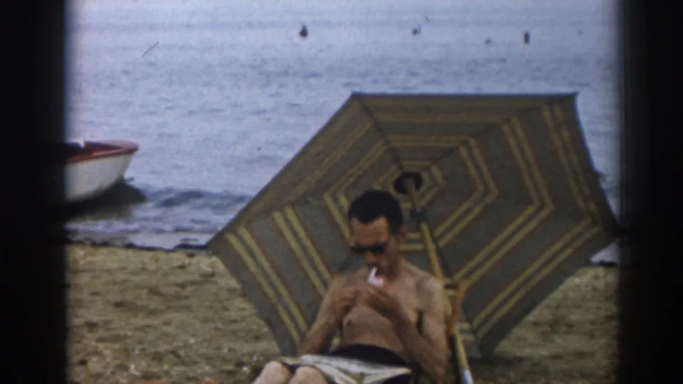 The 1963 beach scene shows the man looking down at reading material while smoking under the umbrella near the shoreline.