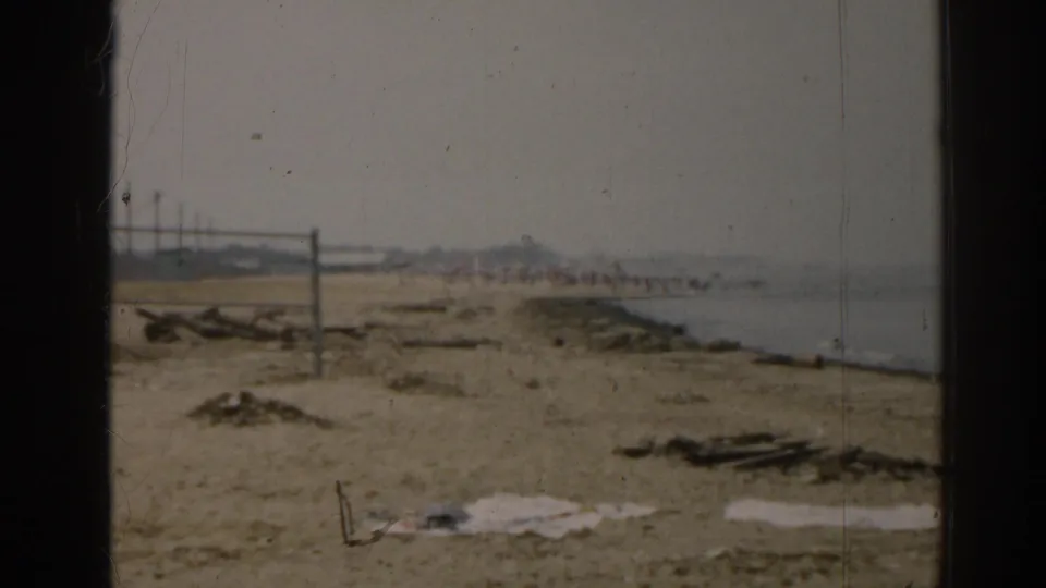 1963 New Jersey beach view on 8mm film, showing sand, shoreline water, scattered beach material, and a distant net.
