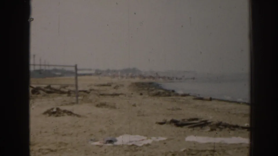 1963 New Jersey shoreline view with a sandy beach, calm water, scattered material, and a distant beach net in the background.