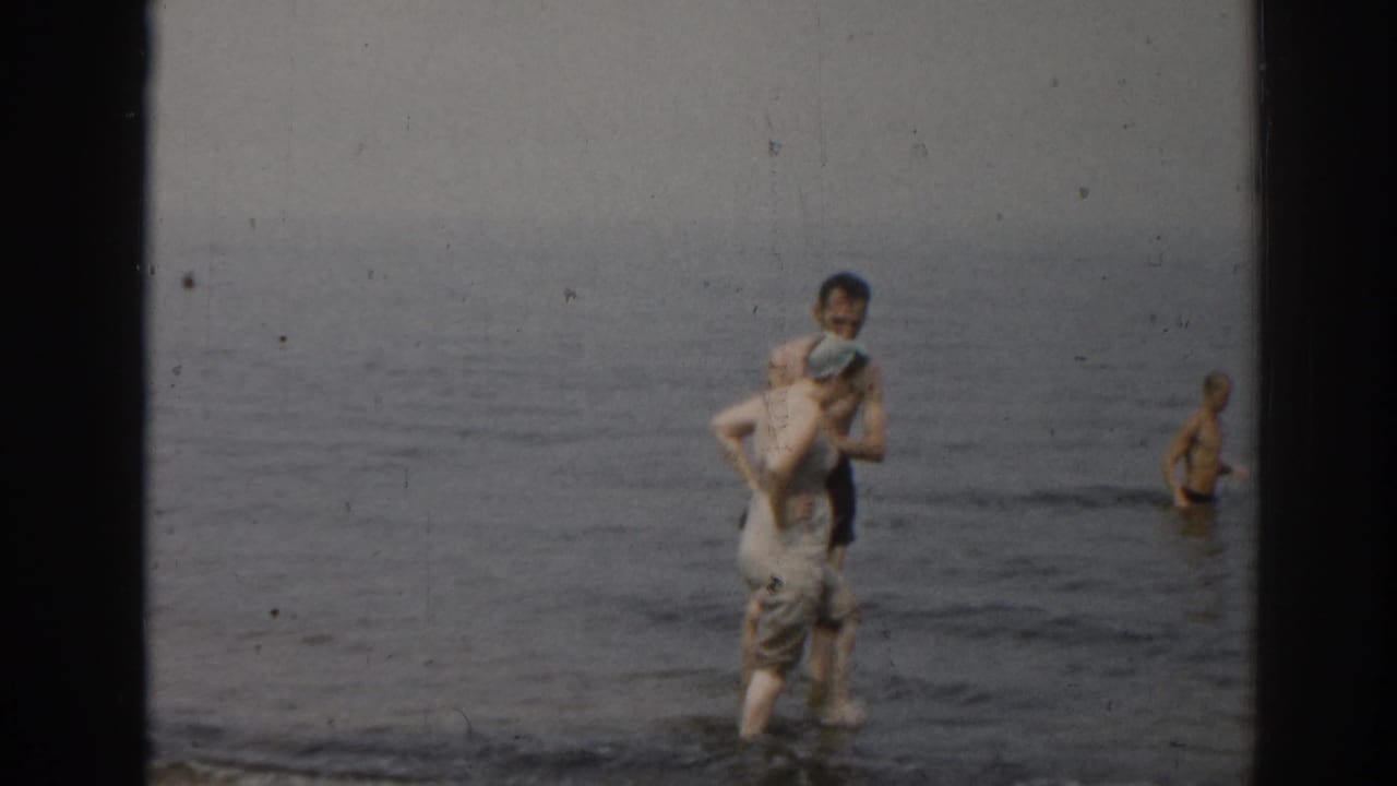 Man in dark swim trunks carries a woman in light clothing through shallow ocean water.