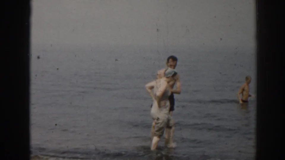 1963 New Jersey home-movie frame of a man carrying a woman in light beach clothing through shallow surf with another bather behind them.