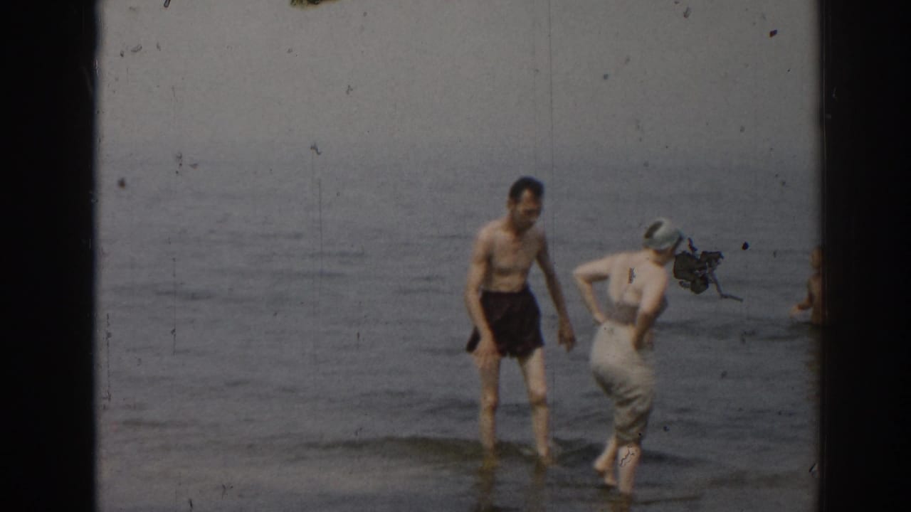 Man and woman stand in shallow surf while another swimmer is partly visible at the frame edge.