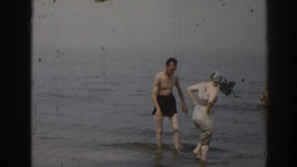 1963 New Jersey beach frame showing a man in dark trunks beside a woman in a swim cap and light clothing at the waterline.