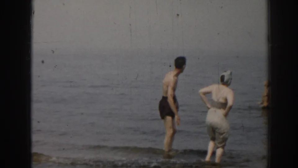1963 New Jersey home-movie frame of two adults from behind in the shallow surf, with the woman wearing a swim cap and light clothing.