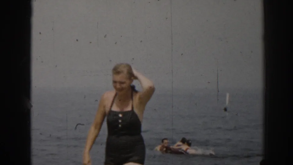 1963 New Jersey beach frame with a woman in a black one-piece swimsuit walking near the camera and bathers floating in the ocean behind her.