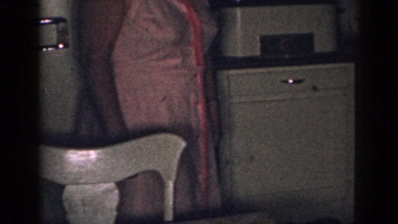 Woman partly visible beside a white chair and refrigerator in a dim kitchen.