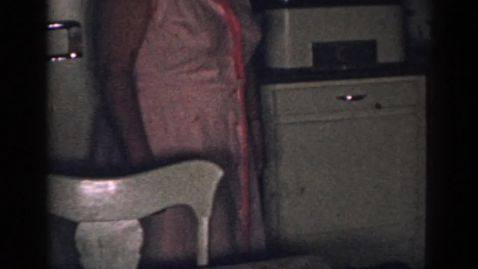 1963 New Jersey home-movie frame showing a woman partly cropped beside a white chair and retro refrigerator in a dim kitchen.