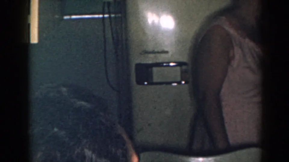 1963 New Jersey frame showing a dim kitchen with refrigerator details, a chair back, and the woman cropped at the right edge.