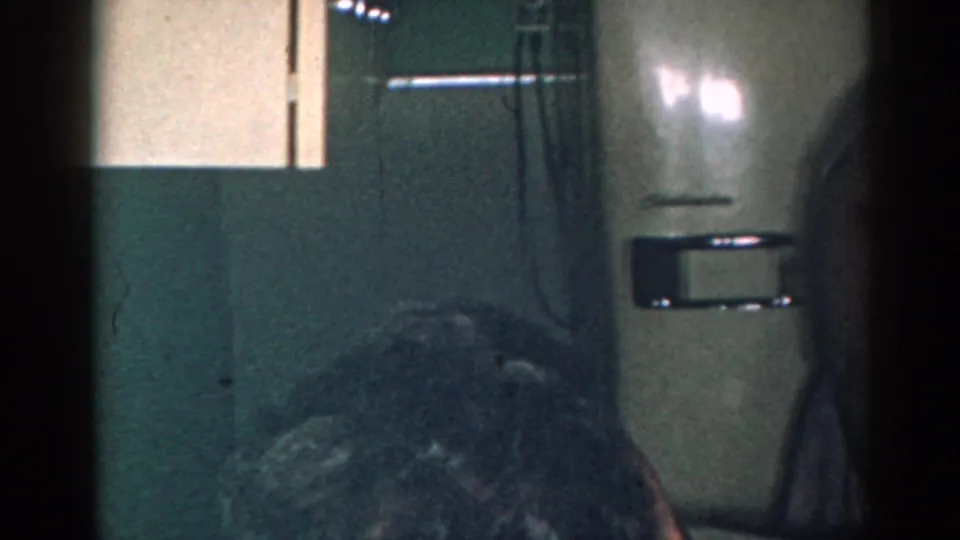 1963 New Jersey home-movie frame of the refrigerator and chair in a dim kitchen, with the woman mostly cropped at the right edge.