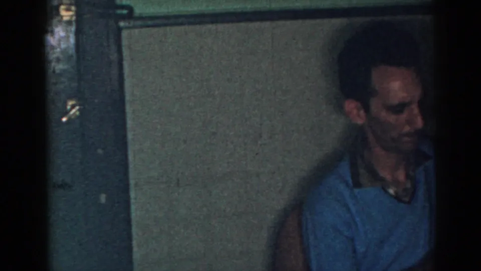 Adult man in a blue sleeveless sweater and collared shirt stands in a dim 1963 New Jersey house interior.