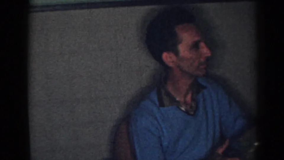 Adult man in profile inside a dim New Jersey home, wearing a blue sweater over a collared shirt.